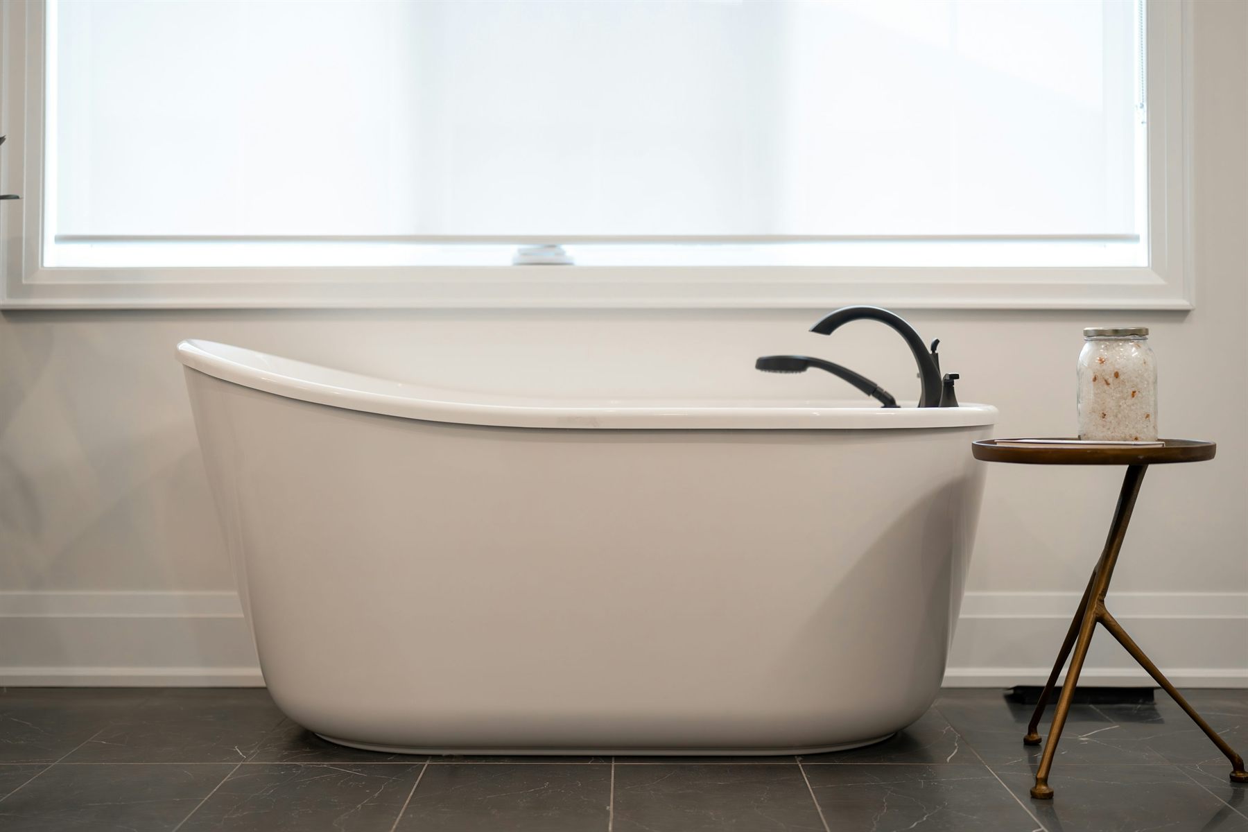 Freestanding white bathtub against a simple wall with dark floor tile and a small side table