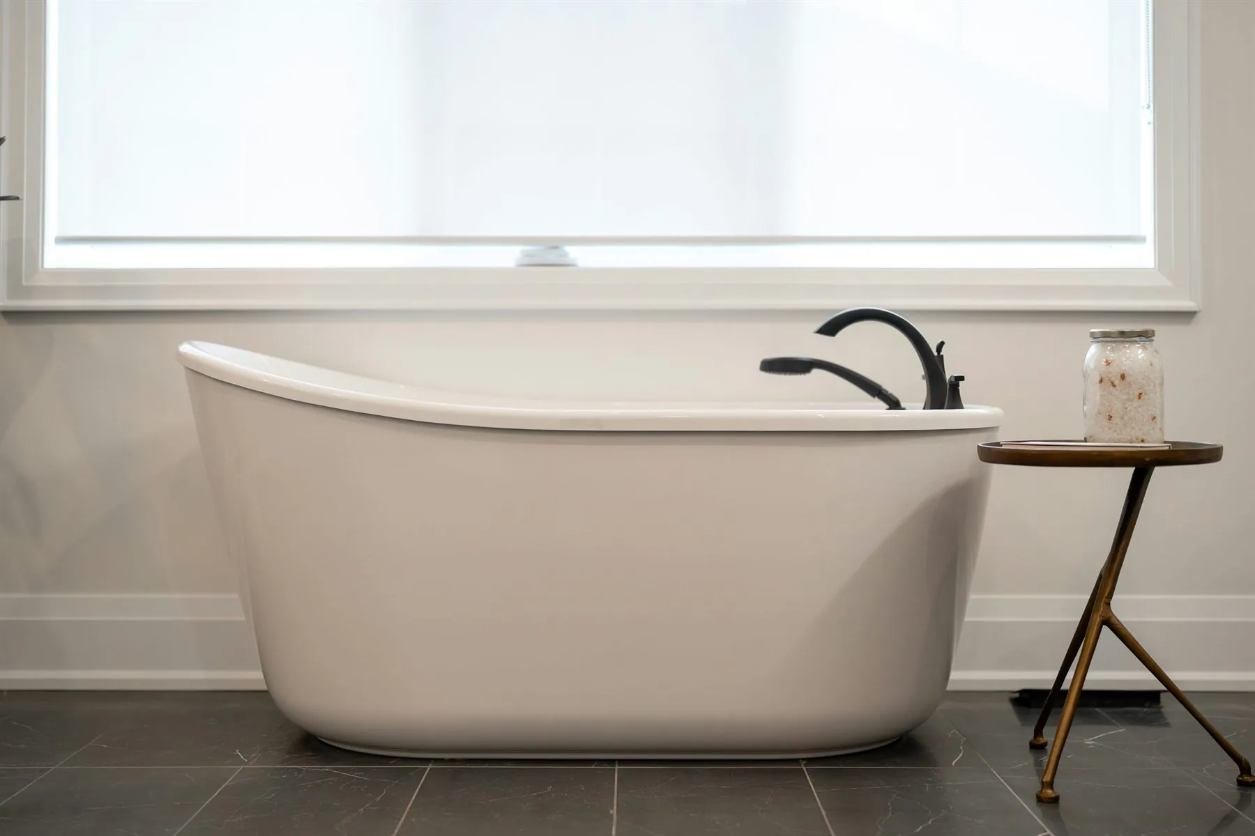 Freestanding white bathtub against a simple wall with dark floor tile and a small side table