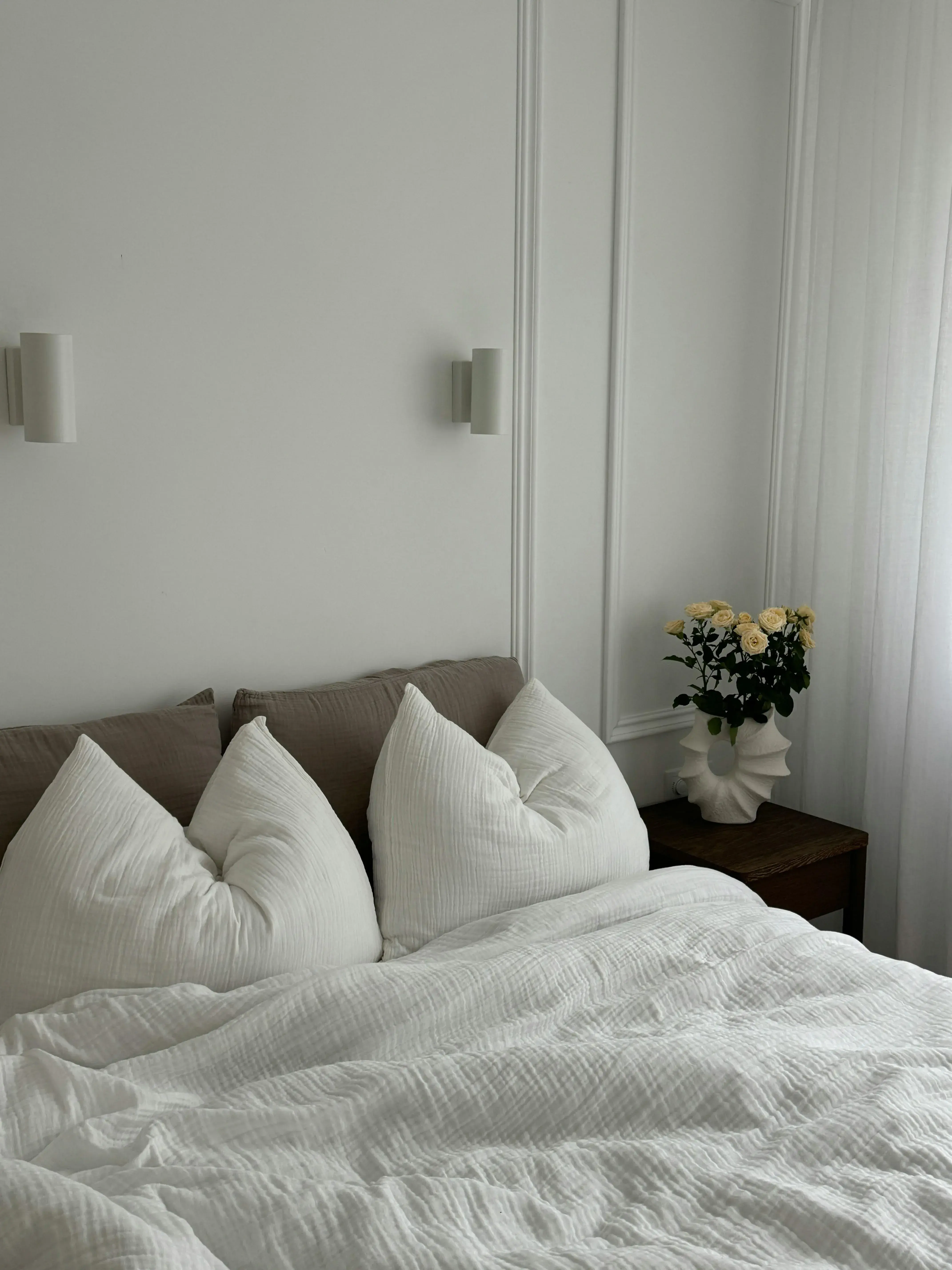 Soft white bedroom corner with one quiet bedside table, flowers, and an edited sleep zone