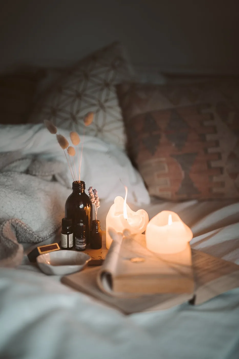 Soft bedroom scene with two lit candles, amber bottles, and an open book on rumpled bedding