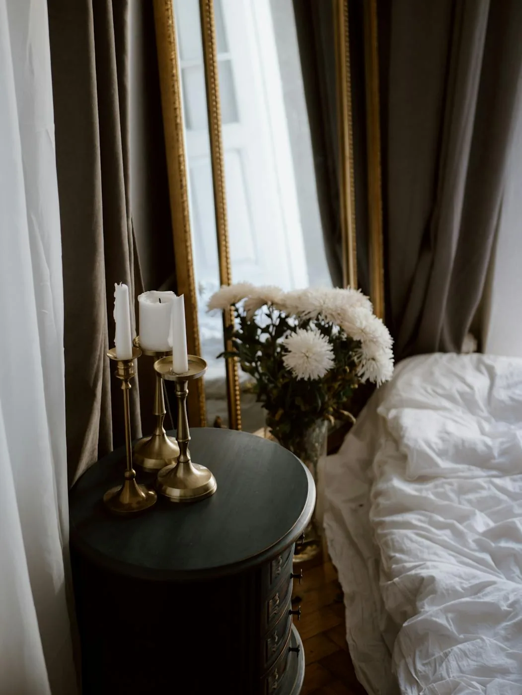 Two white taper candles in brass holders on a dark round bedside table beside a mirror and flowers