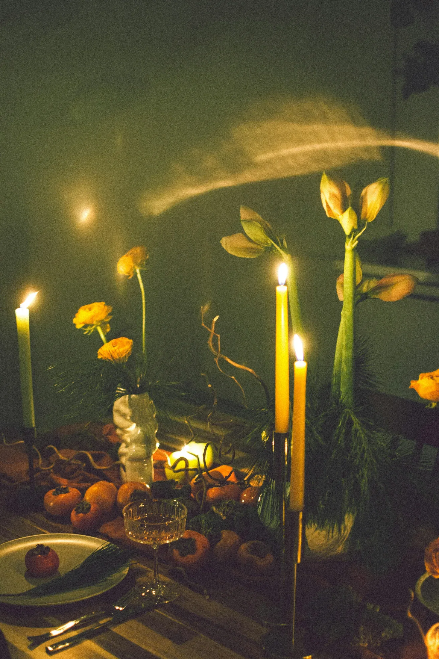 Tall lit candles on a warm dining table with soft yellow flowers in the background