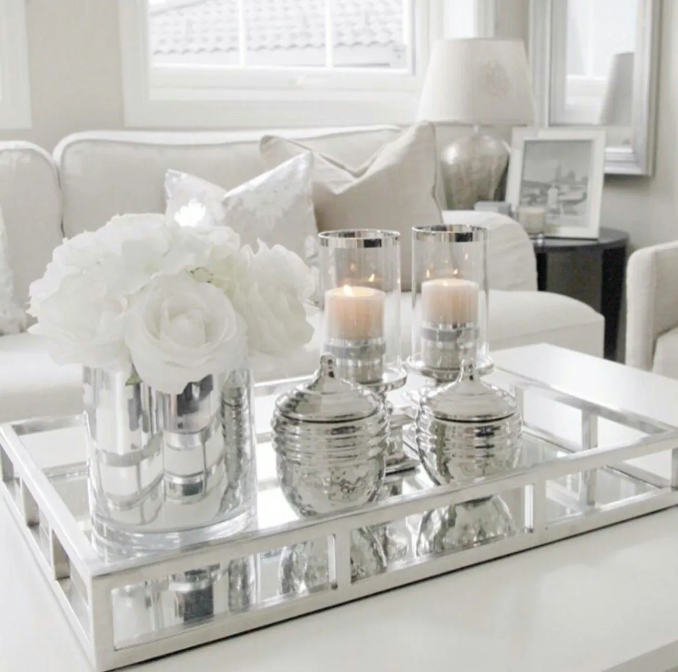Neutral glass candle holders arranged on a reflective tray with soft white flowers nearby