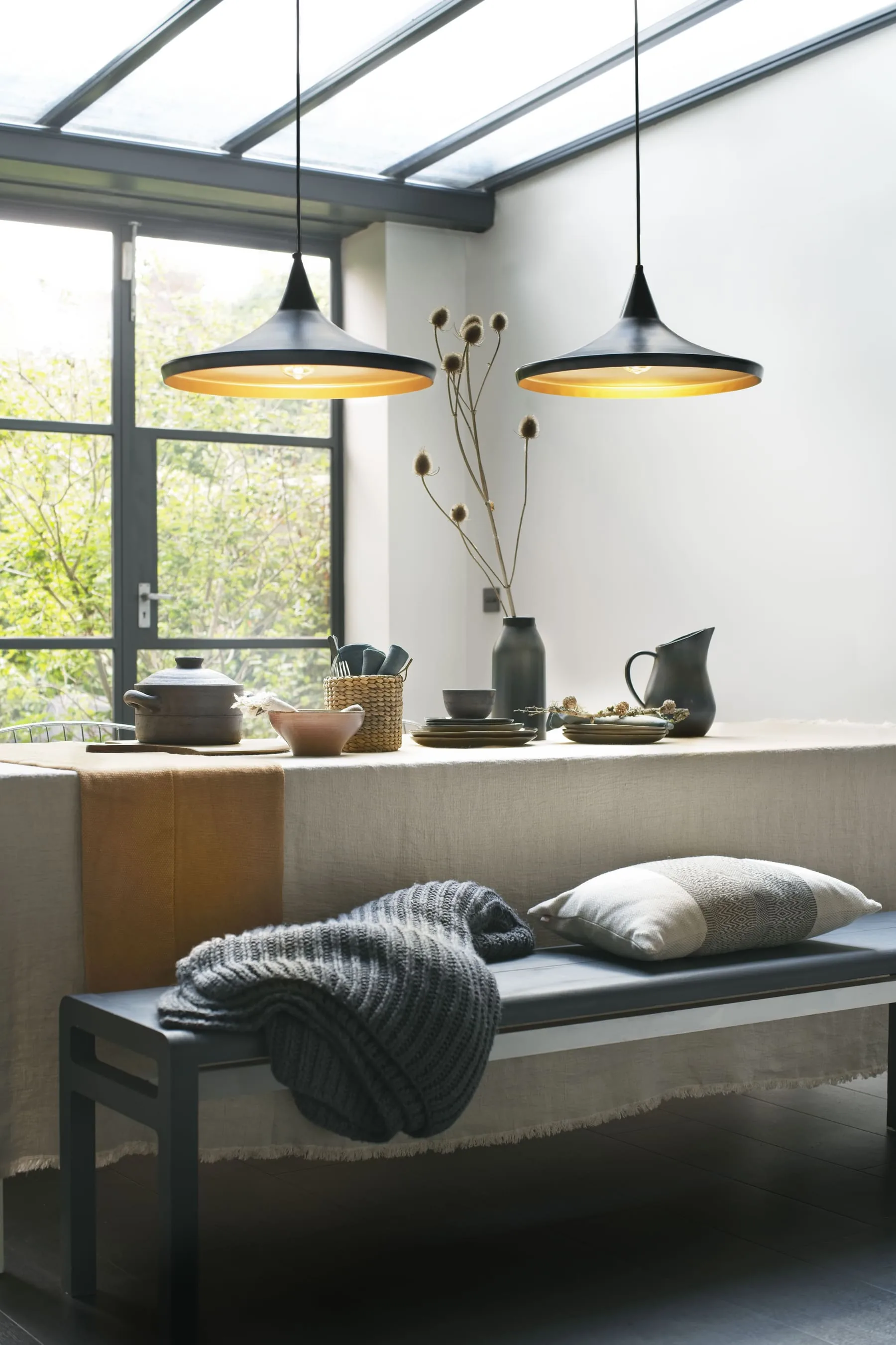 Dining table under two black pendant lights with a bench, textured textiles, and a softer neutral setting