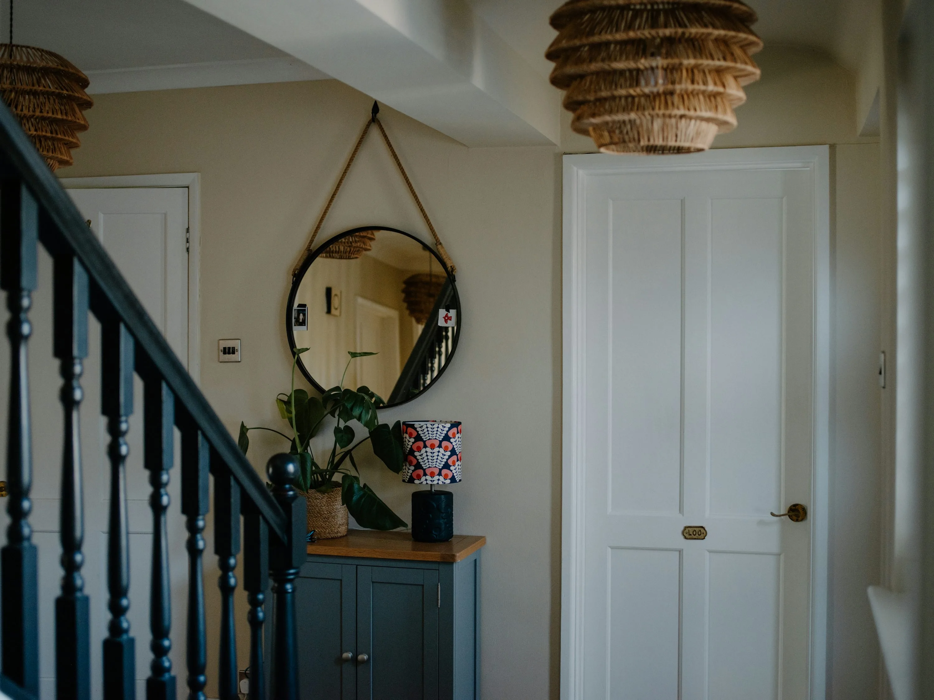 Entryway corner with a small cabinet, round mirror, lamp, plant, and nearby staircase