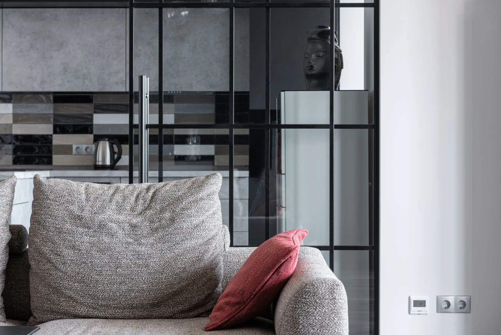 A muted red cushion on a textured gray sofa beside a glass partition and kitchen