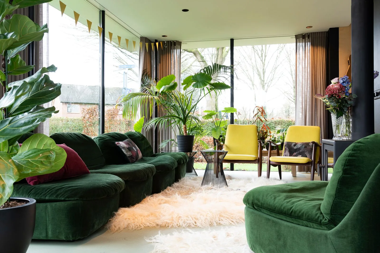 Green sofas and leafy plants beside large windows, with yellow chairs and metal side tables