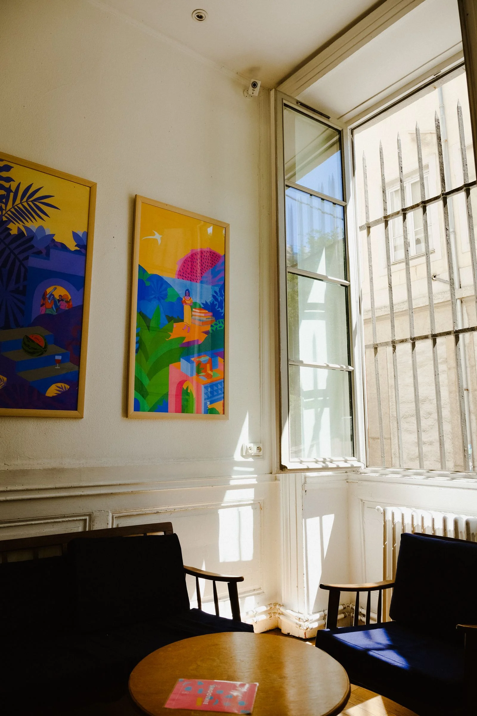 Two colorful yellow and blue framed prints beside a window and dark blue upholstered chairs
