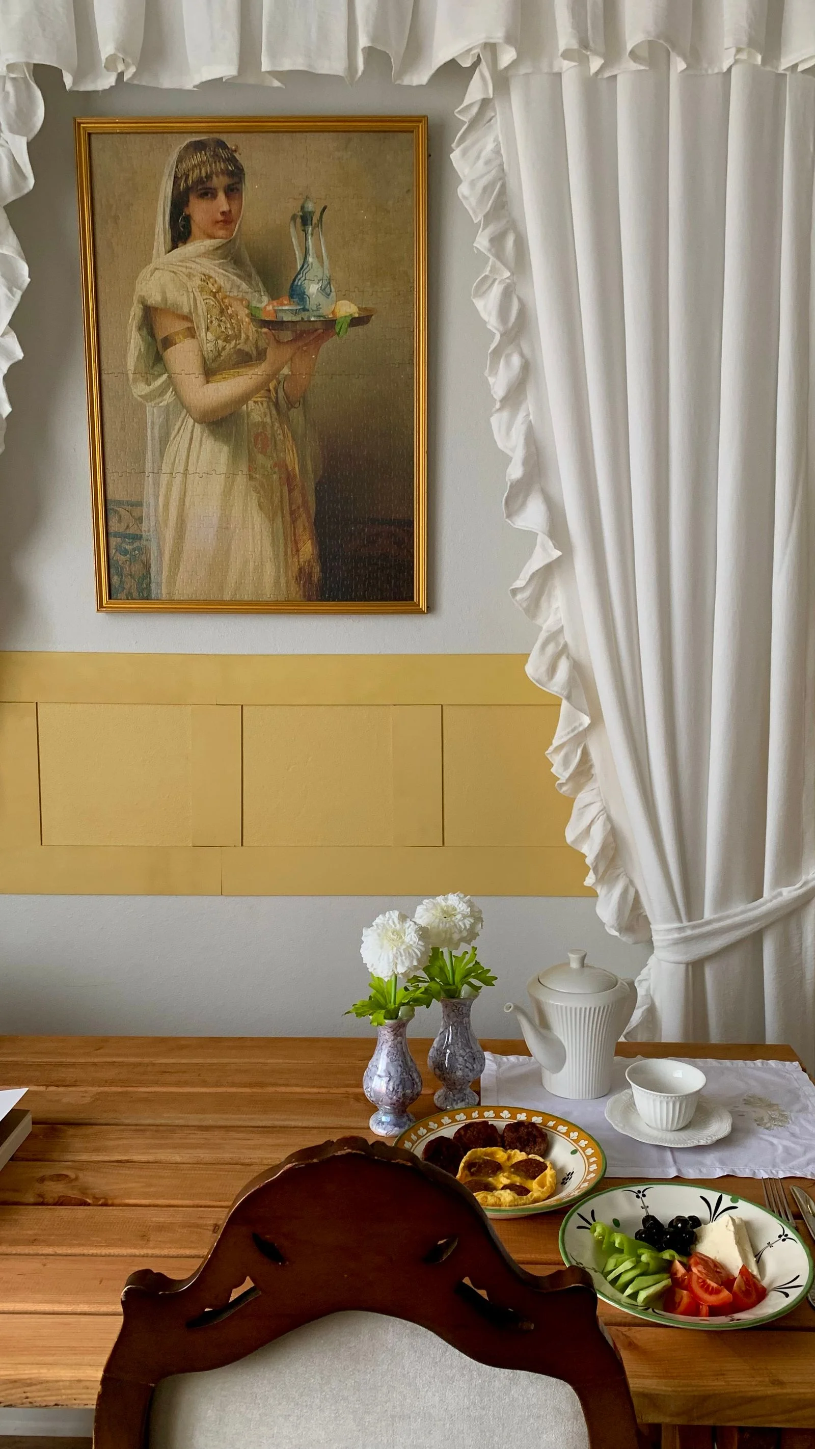 Framed portrait above a wooden dining table beside white curtains