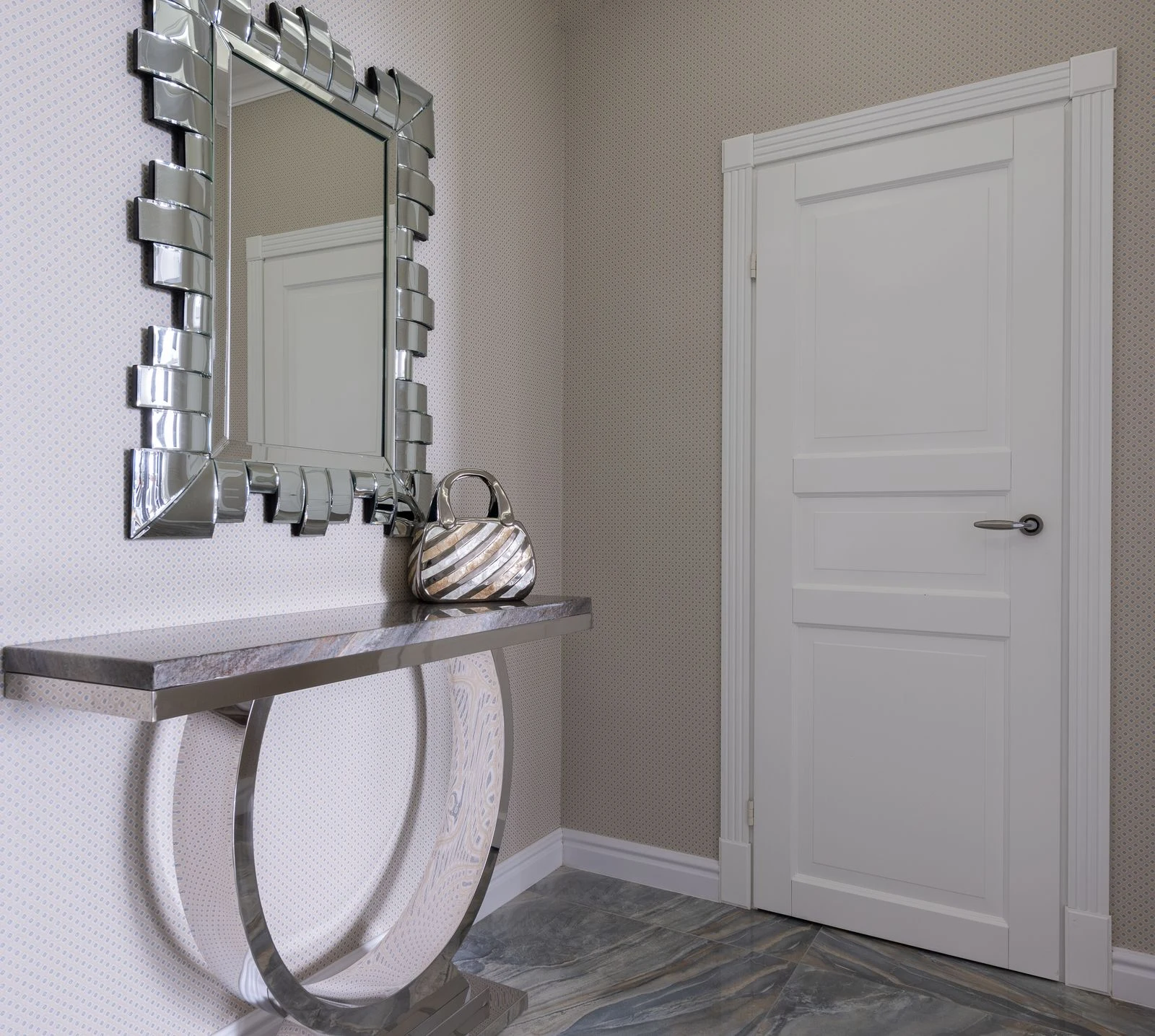 Narrow console holding a handbag beside a white door, with a decorative silver-framed mirror above it