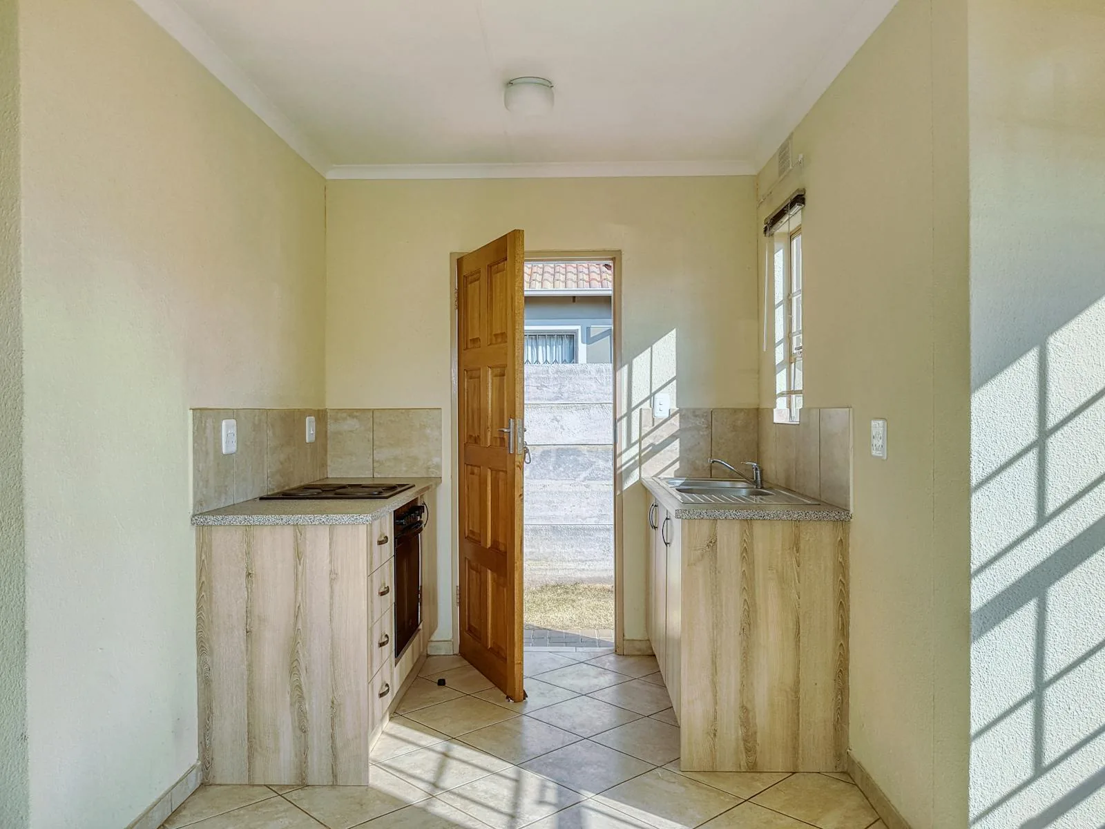 Simple kitchen with pale cabinets, open door, and strong daylight across the floor