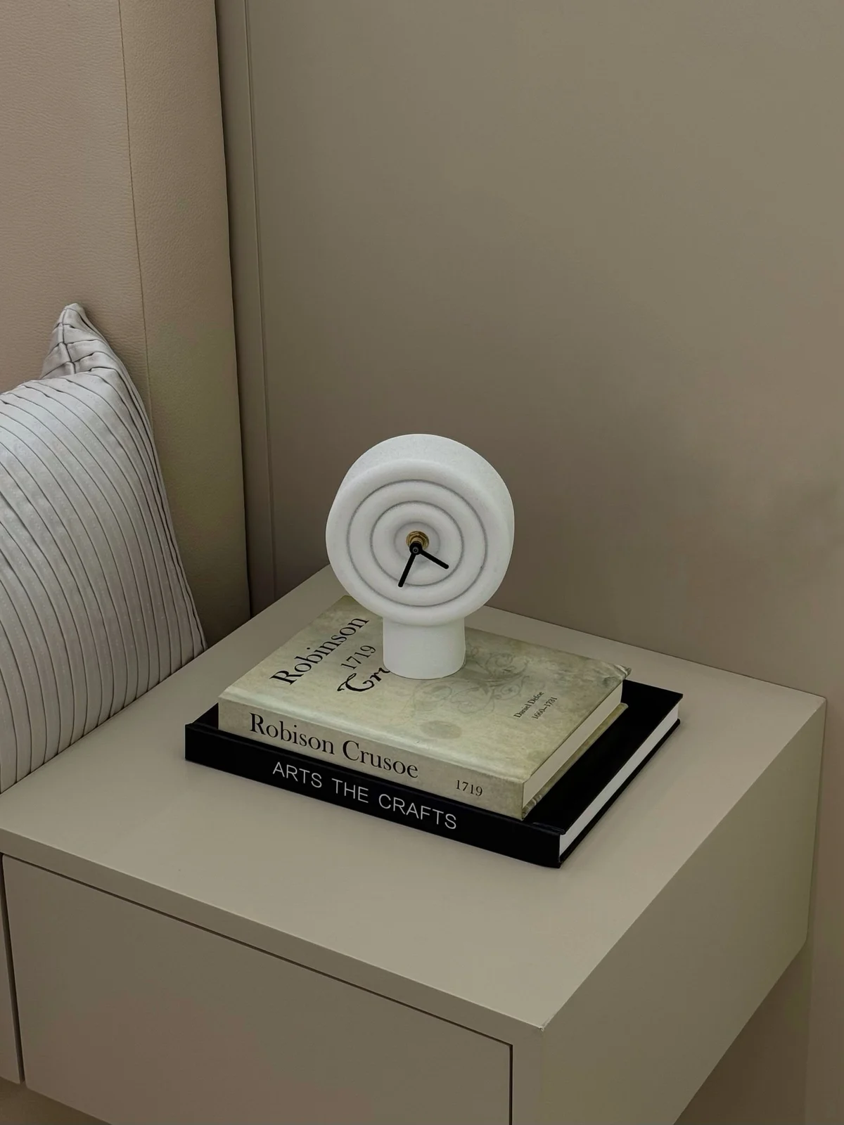 Small white sculptural analogue clock on books beside a bed