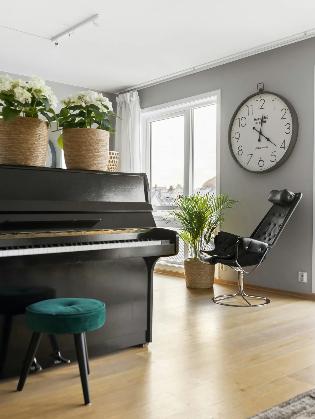 Large wall clock beside a window above an armchair and near a piano