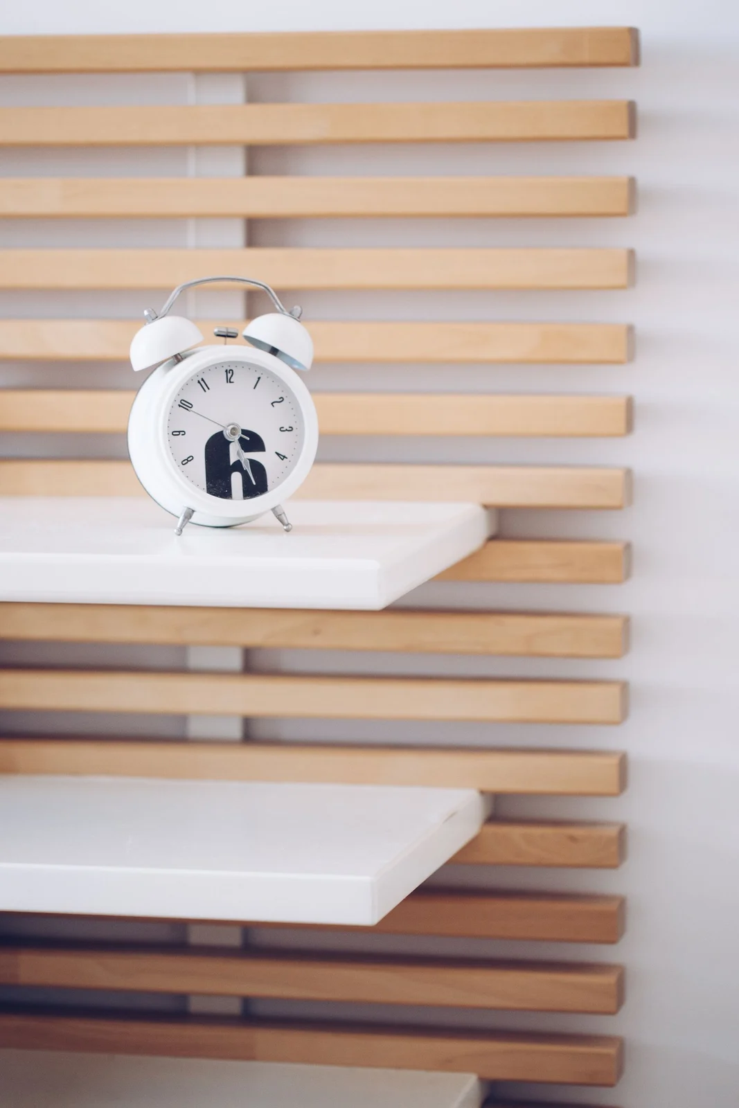 White analogue alarm clock resting on a small white shelf against timber wall slats