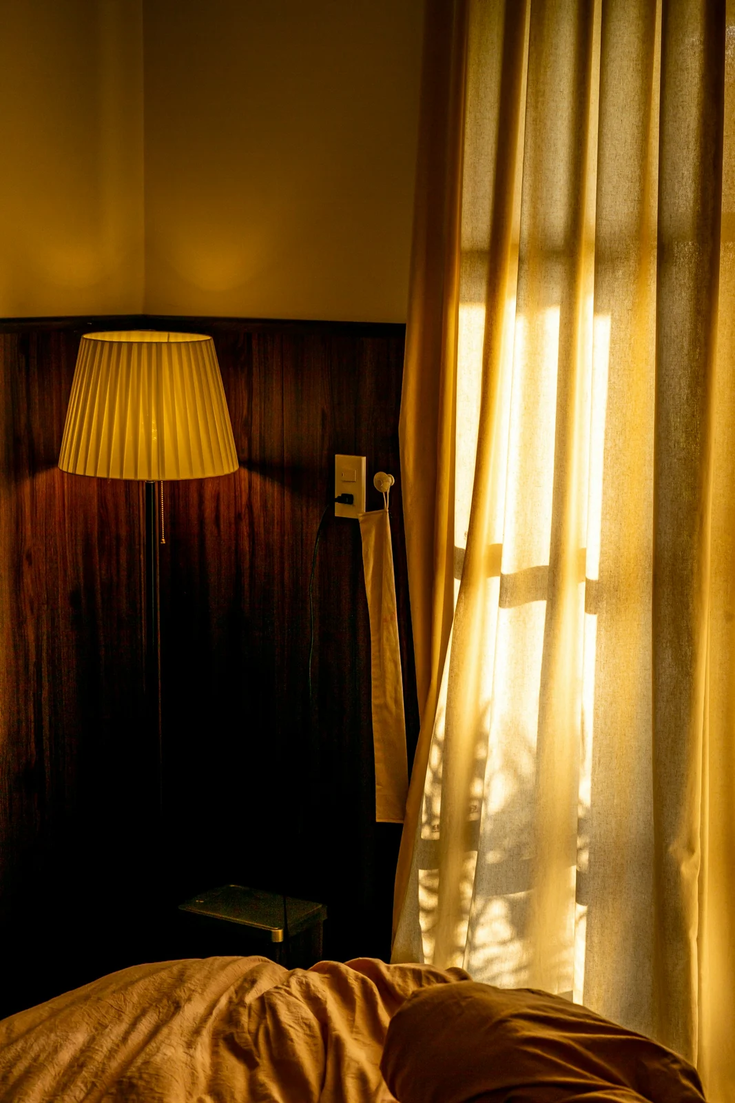 Sunlight passing through a sheer curtain beside a bed and floor lamp