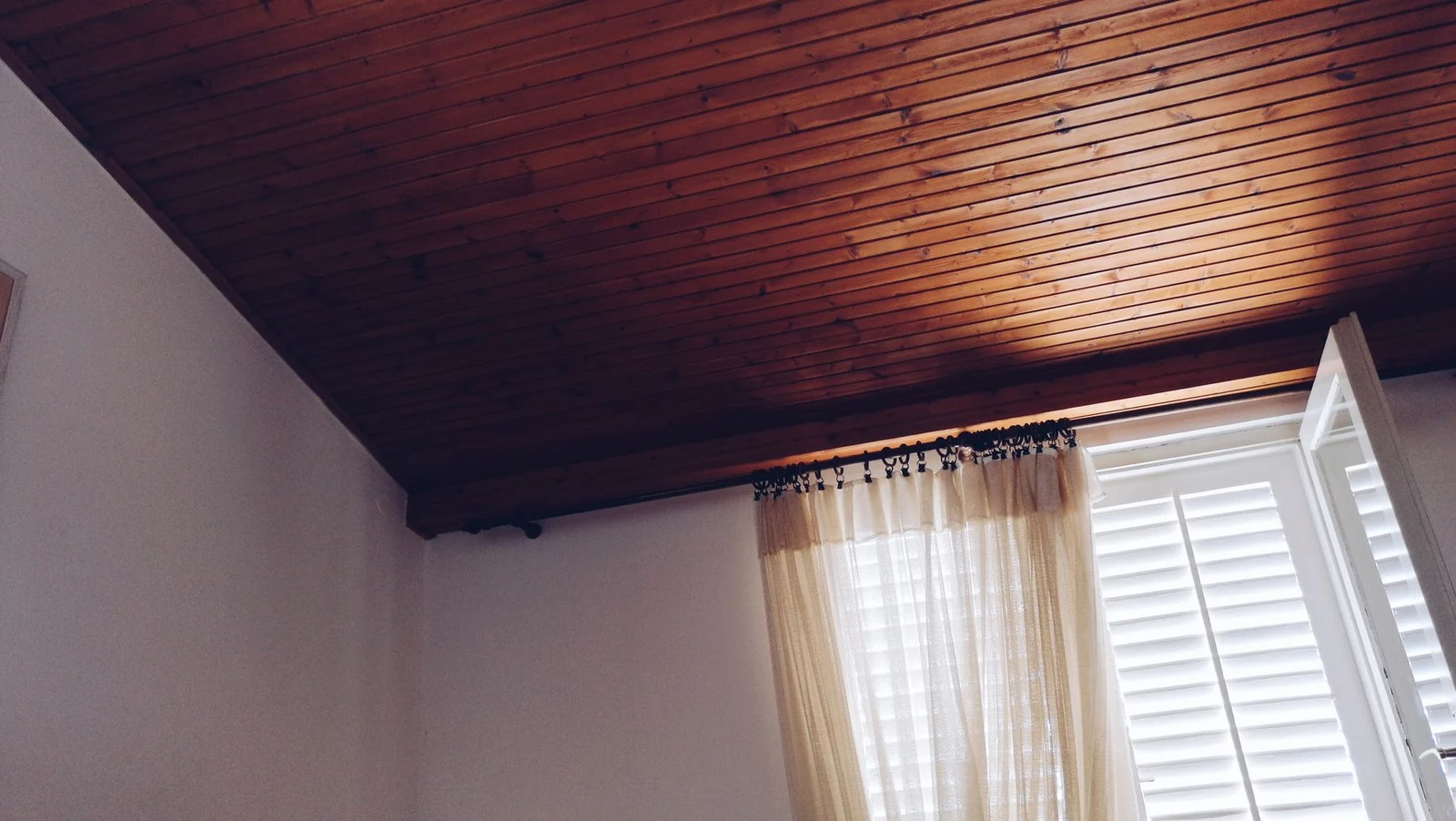 Curtain rings and a sheer panel beneath a timber ceiling beside an open shutter