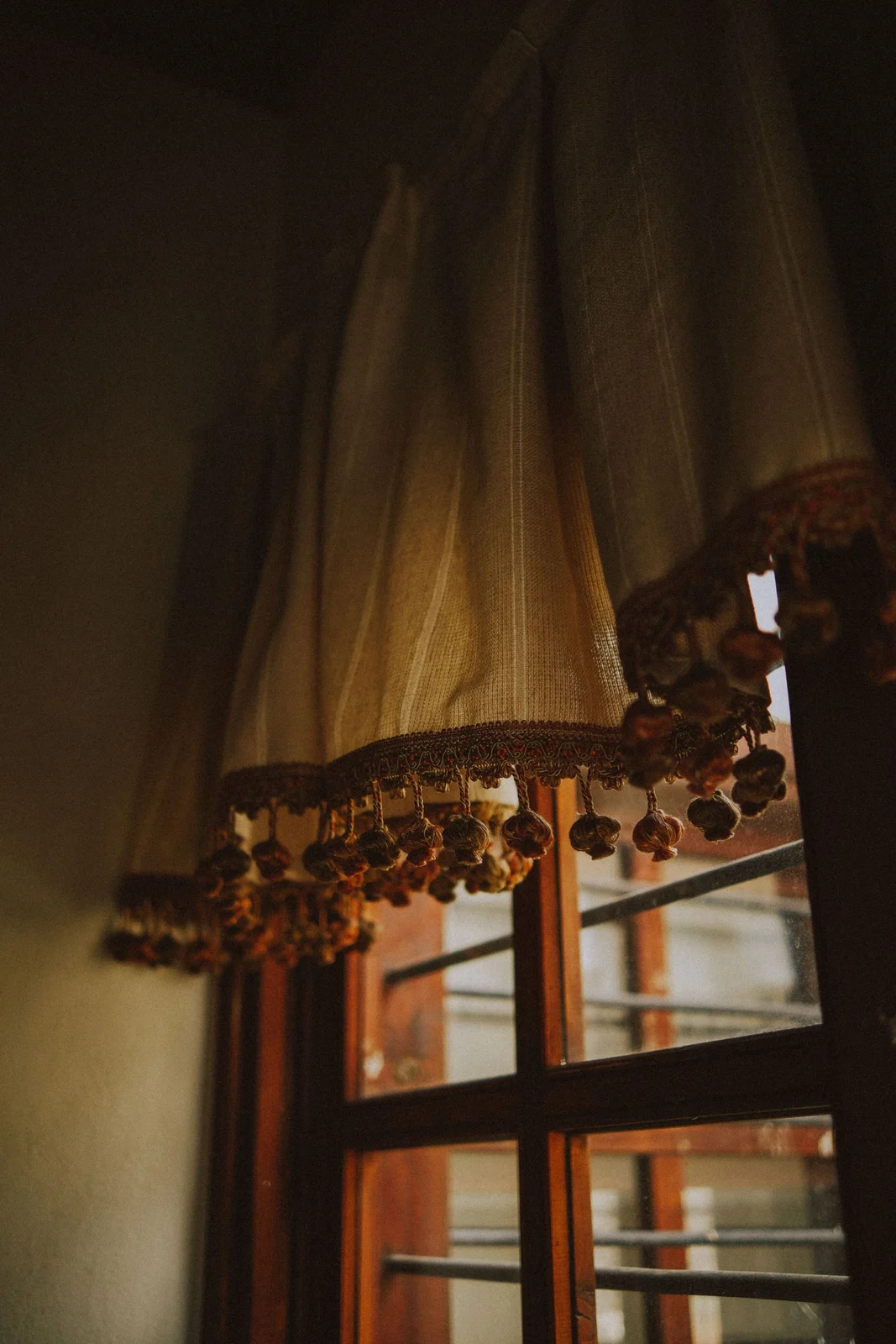 Close view of a light curtain hem with decorative tassels beside a timber window