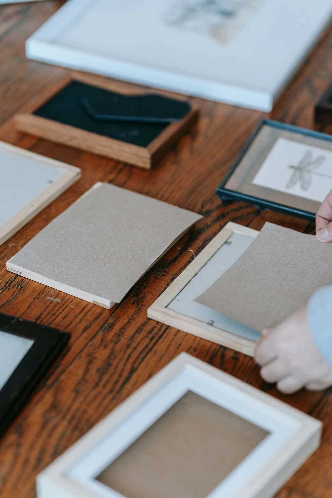 Hands fitting a backing board into a frame among several picture frames on a wooden table