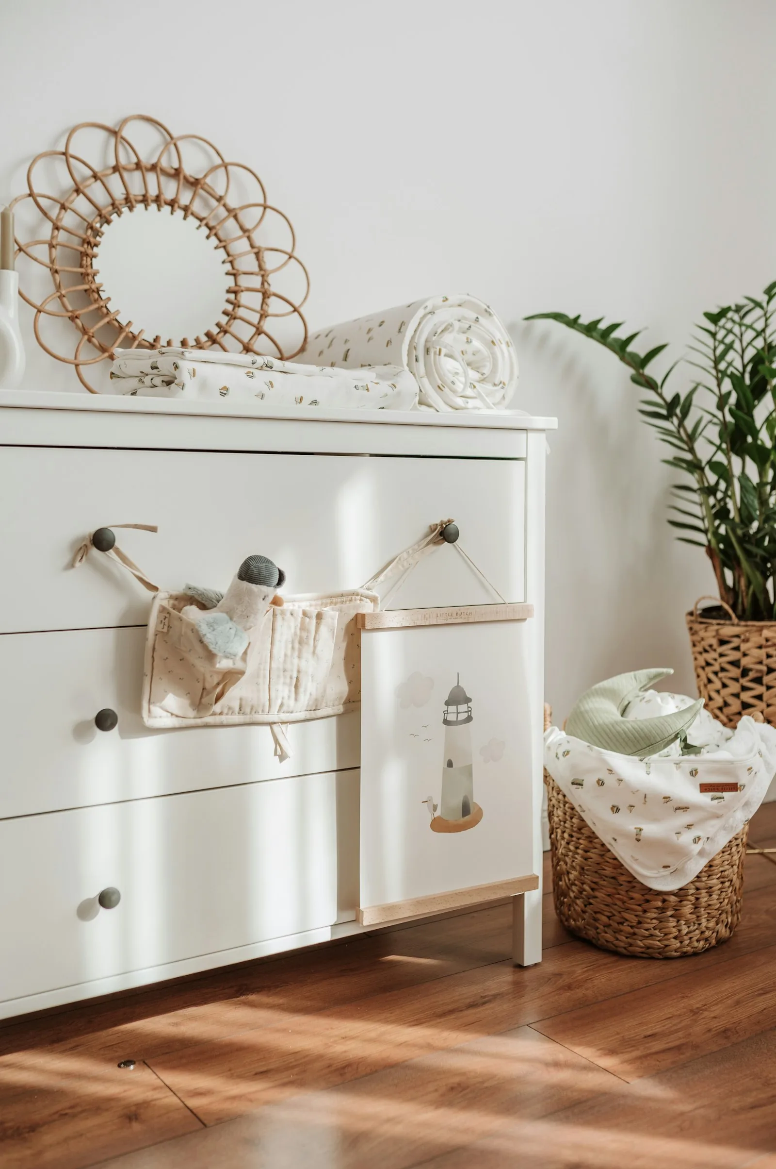 Nursery dresser with folded blankets, a small mirror, hanging organizer, basket, and a plant in soft daylight