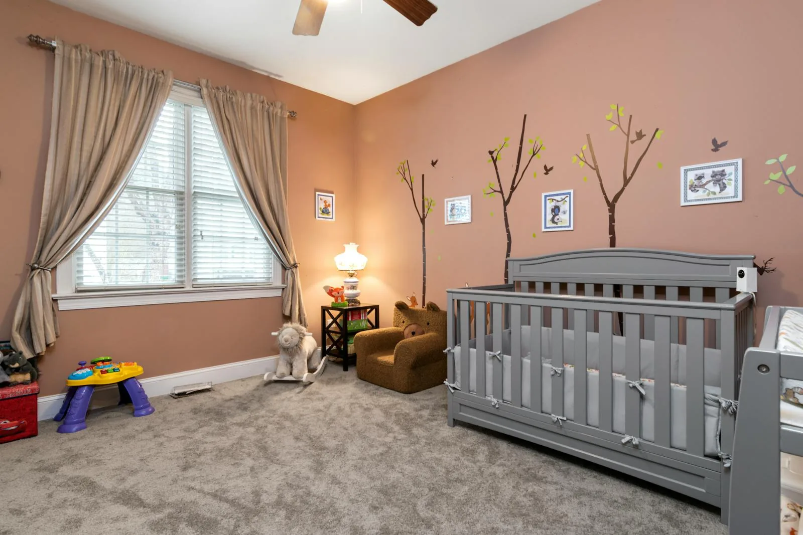 Nursery with a gray crib, warm rose-beige walls, curtains, and one small lamp beside a toddler chair