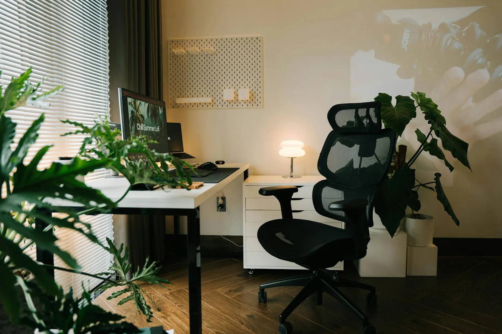 Modern home office with black chair, plants, warm lamp, and soft neutral wall