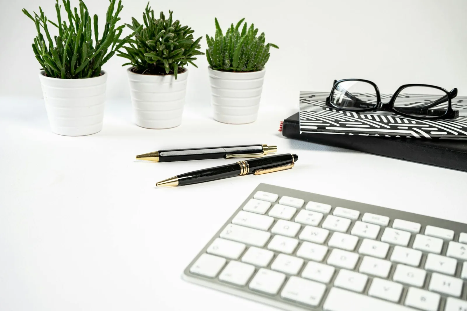 Clean desk with small plants, keyboard, notebook, pens, and white office surface