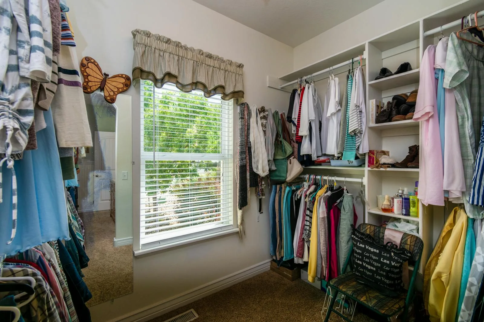 Open closet with hanging clothes, shelves, a window, and a mirror beside the wardrobe