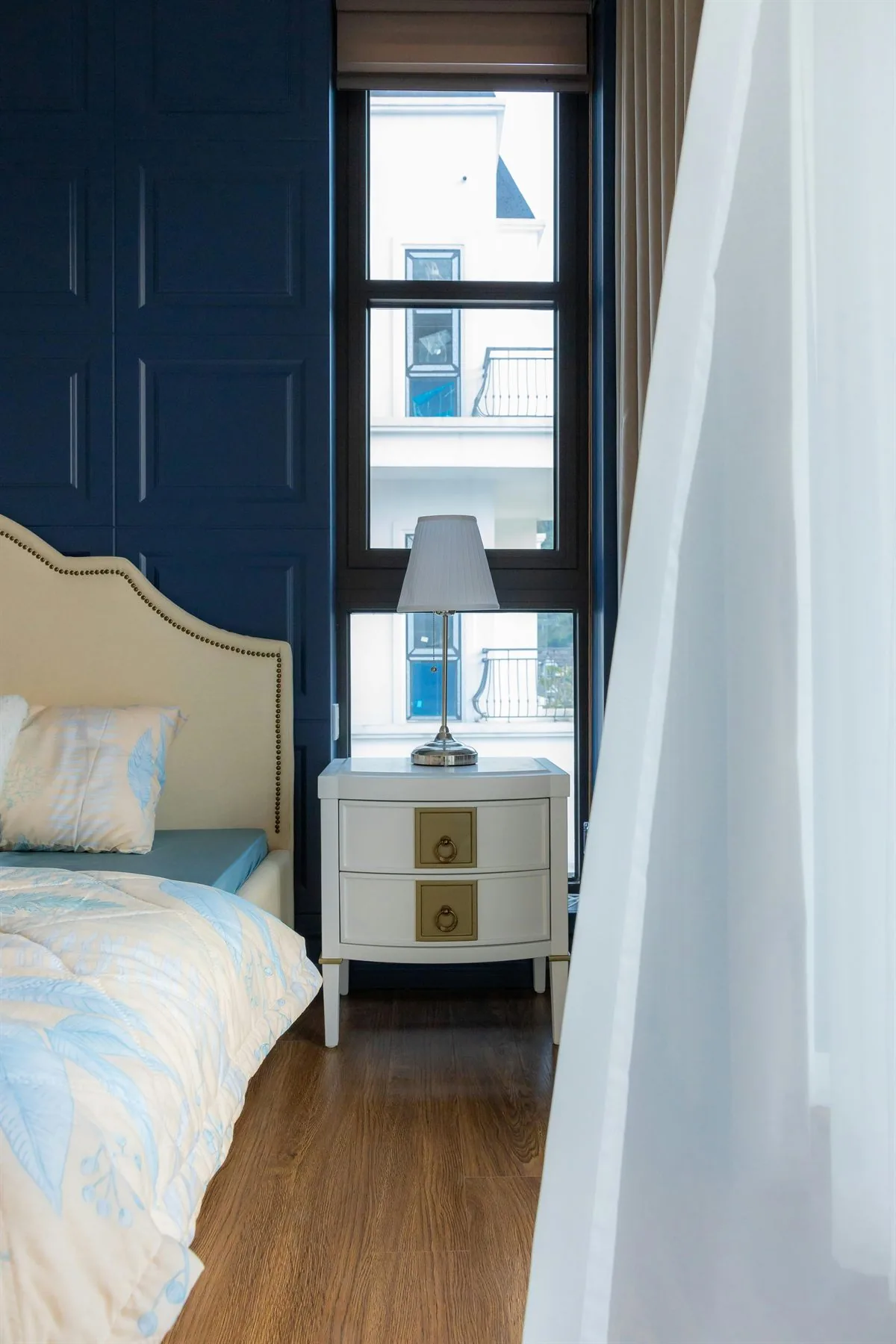 Bedroom with deep blue wall, pale bedding, bedside lamp, and soft curtains